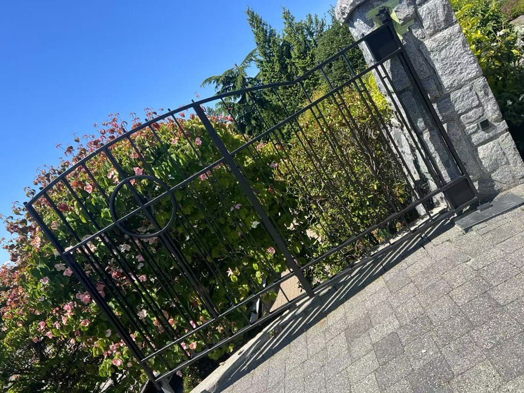 A black wrought-iron gate partially open, set against a backdrop of lush green shrubs and trees under a clear blue sky
