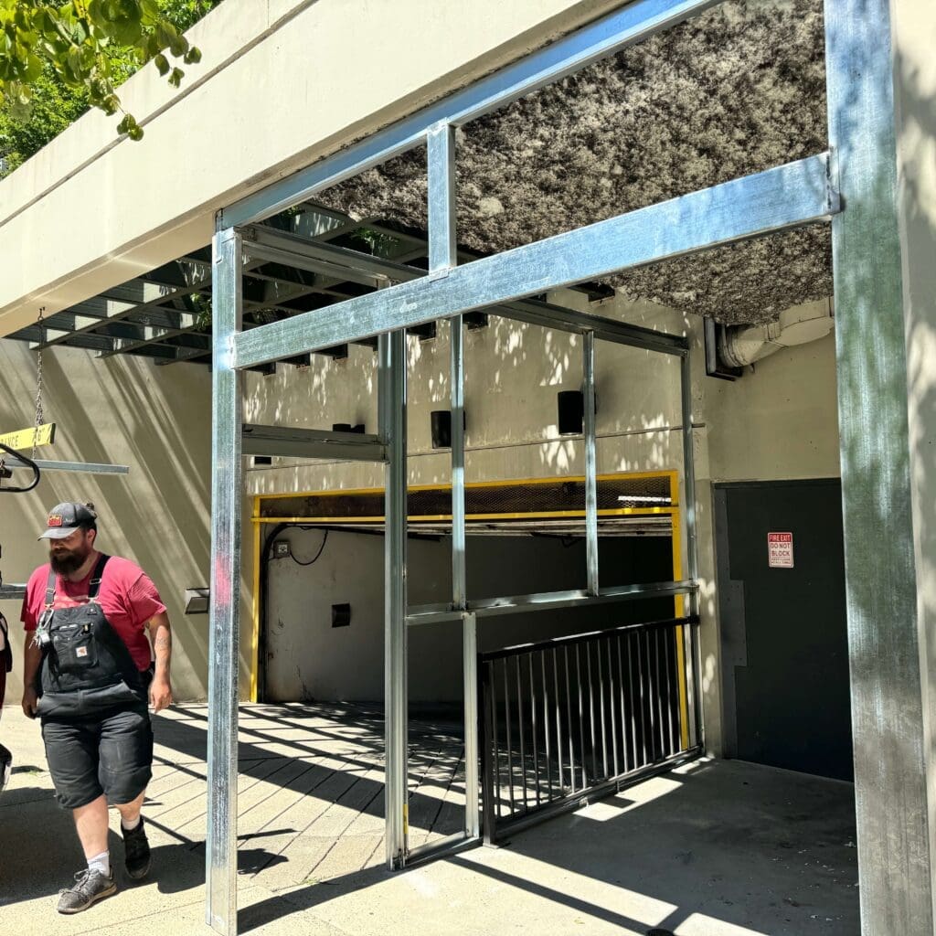 A man in a red shirt and black hat walks along a walkway under a partially constructed building, with metal framing visible above him and greenery in the background.