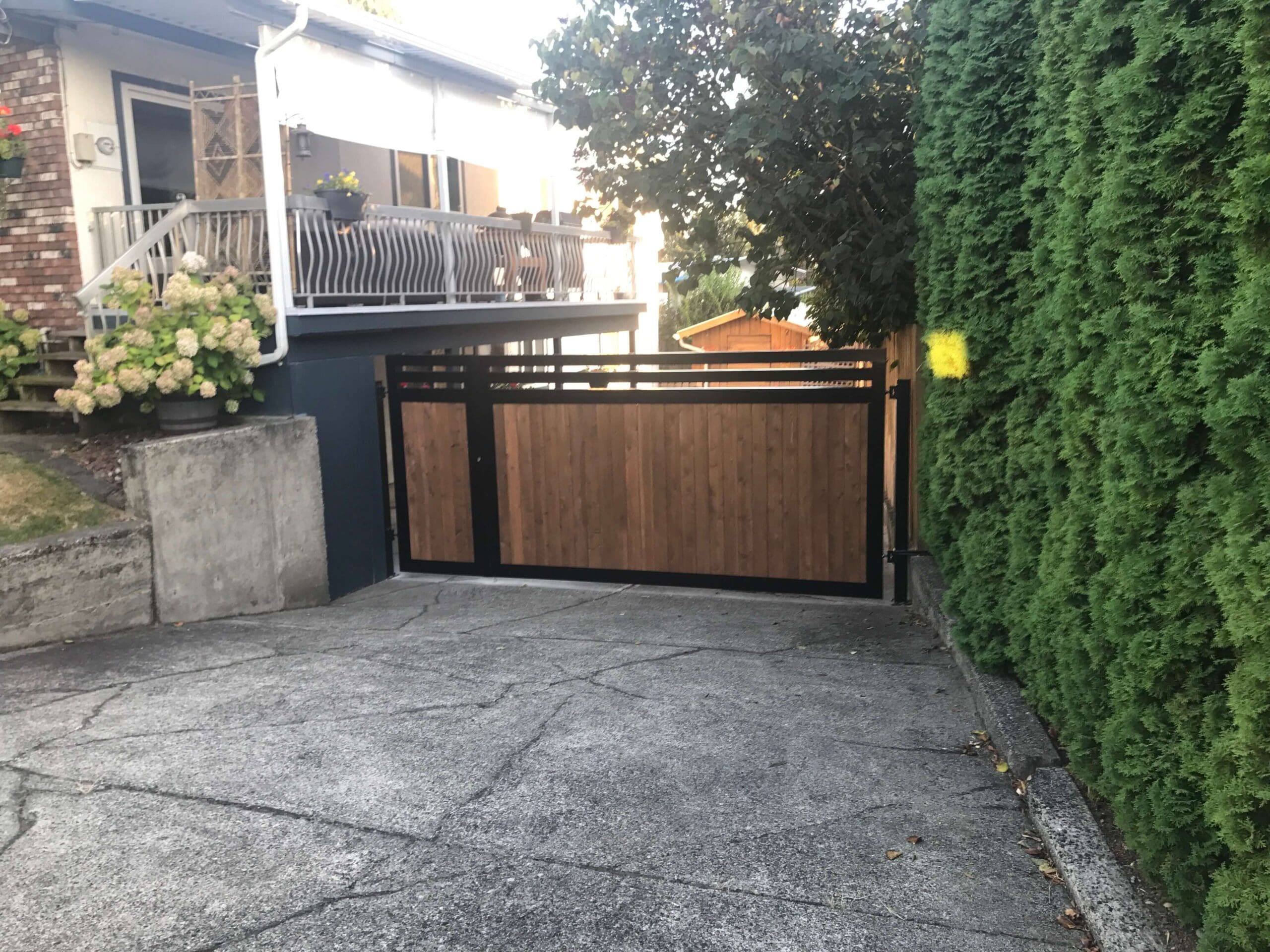 A wooden and metal driveway gate is closed at the end of a concrete driveway, with a house, plants, and a hedge visible nearby