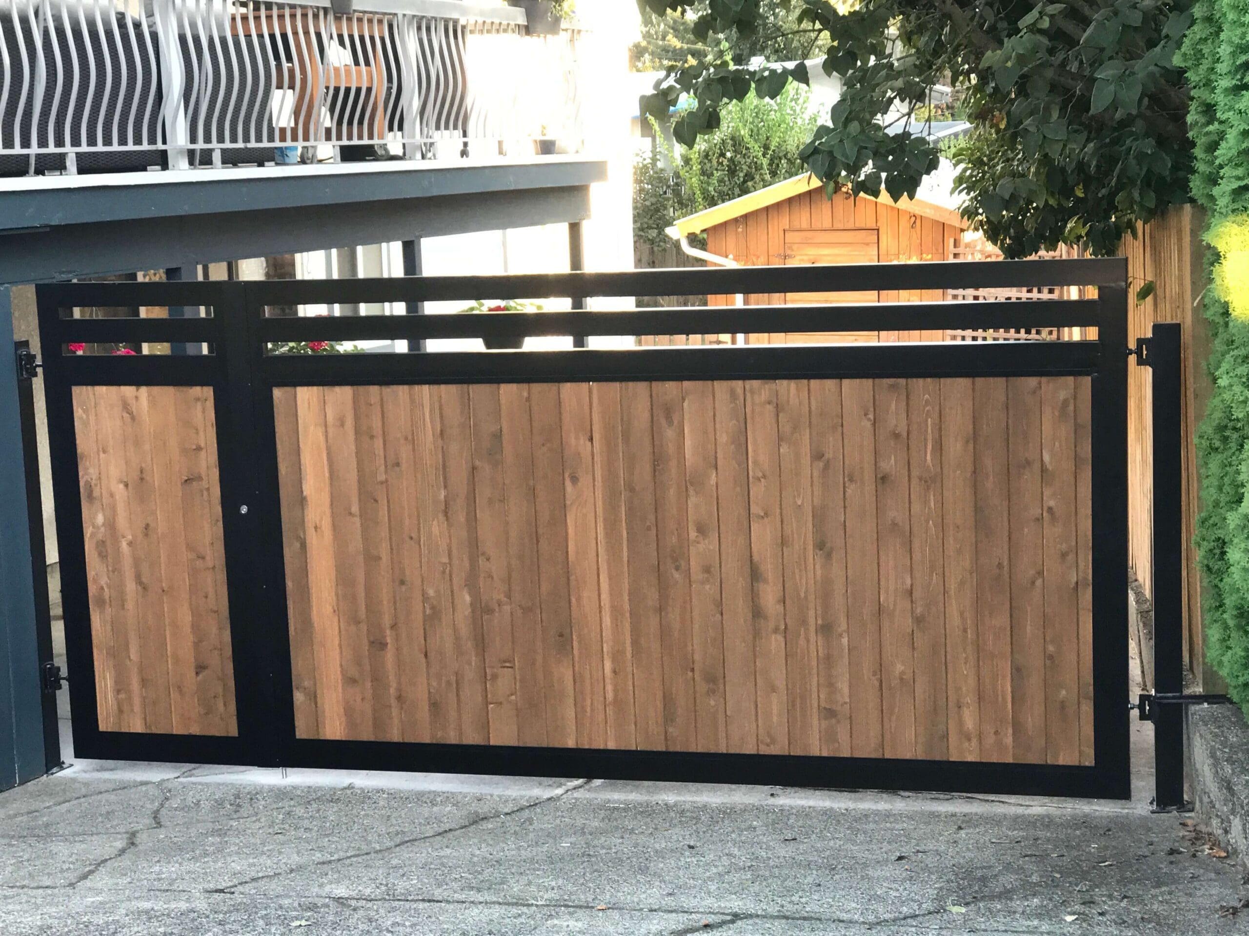 A modern wooden gate with a black metal frame, partially open, leading to a driveway