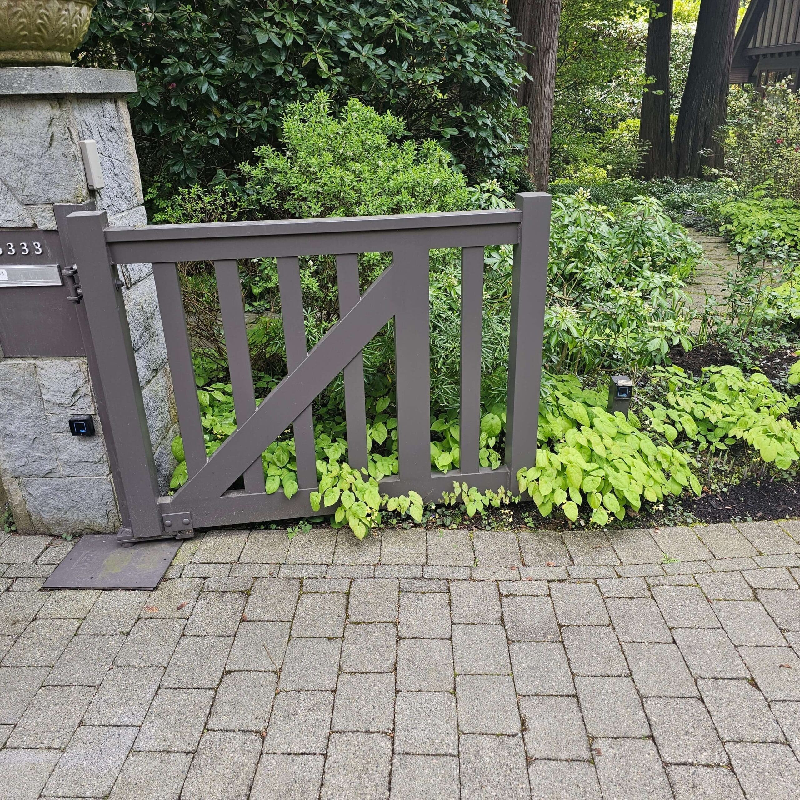 A gray wooden gate is partially open, framed by lush green plants and foliage, set on a stone wall with a paved pathway in front.