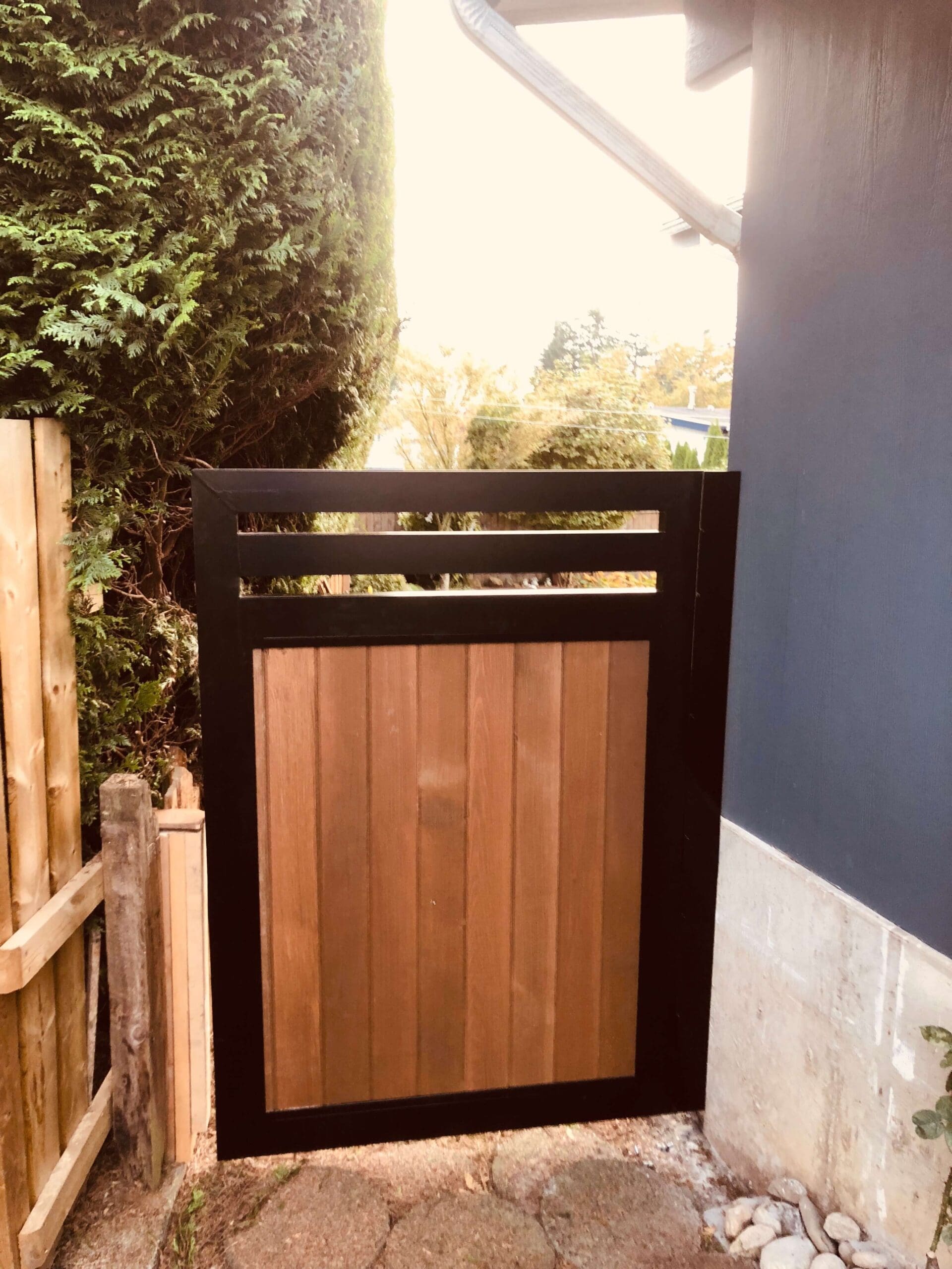 A wooden gate with a black frame, standing open next to a blue wall, revealing a path lined with greenery.